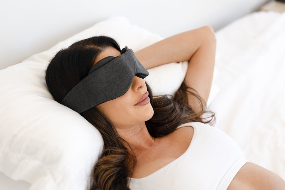 Woman wearing a blackout sleep mask lying on a white pillow.